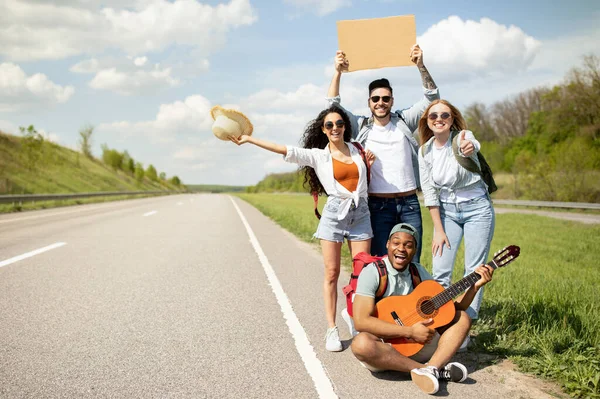 Joyful amigos multiétnicos batendo o seu passeio na estrada com sinal vazio, tocando guitarra, indo em viagem autostop — Fotografia de Stock