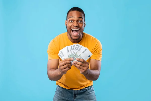 Homem negro excitado segurando o dinheiro do euro gritando emocionalmente, fundo azul — Fotografia de Stock