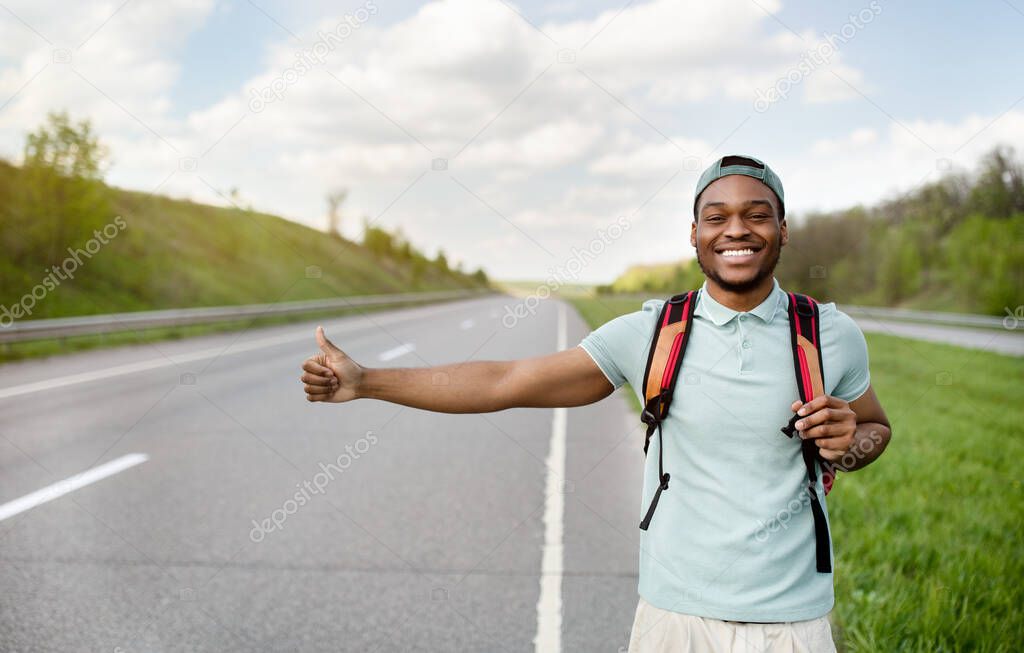 Smiling black guy with backpack flagging down car, catching free ride, hitchhiking on highway, free space