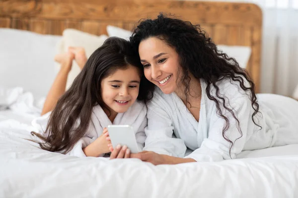 Веселая мать и дочь в халатах с помощью смартфона — стоковое фото