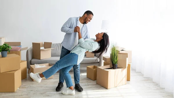 Gelukkig liefdevol Afrikaans Amerikaans paar dansen in hun nieuwe huis tussen kartonnen dozen op bewegende dag, panorama — Stockfoto