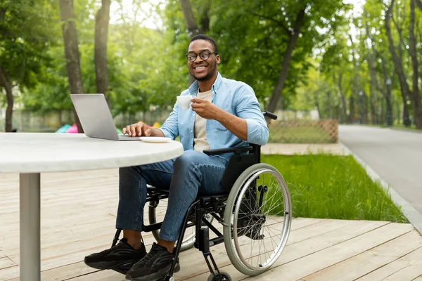 Trabalho remoto para pessoas com deficiência. Joyful cara preto em cadeira de rodas trabalhando on-line, usando laptop no café ao ar livre — Fotografia de Stock