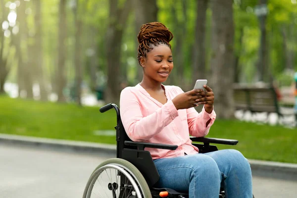 Mujer negra discapacitada feliz en silla de ruedas usando teléfono inteligente, revisando mensajes, navegando por Internet en el parque de la ciudad — Foto de Stock