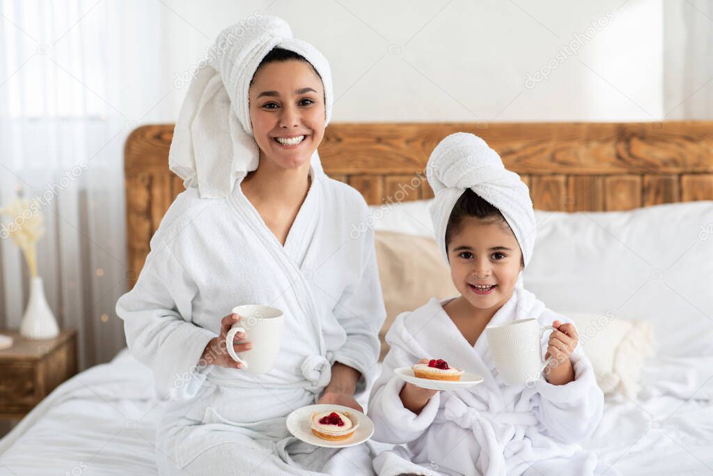 Happy mother and daughter in bathrobes drinking tea with cakes