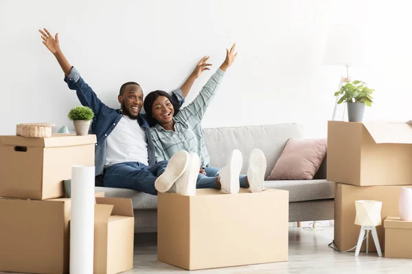 Housewarming Party. Joyful Black Couple Sitting Among Cardboard Boxes And Raising Hands — Stock Photo, Image