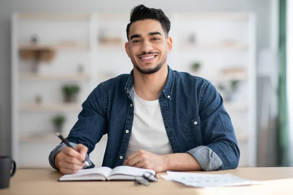 Chico árabe alegre en informal sentado en el escritorio, tomando notas — Foto de Stock