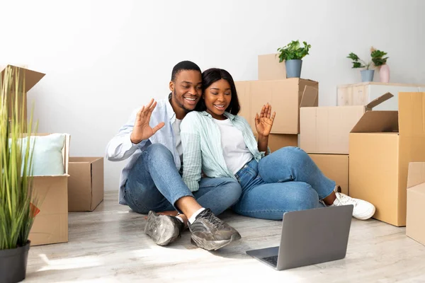 Loving zwart paar met video oproep op laptop computer en zwaaien naar de camera, zitten op de vloer in hun nieuwe huis. — Stockfoto