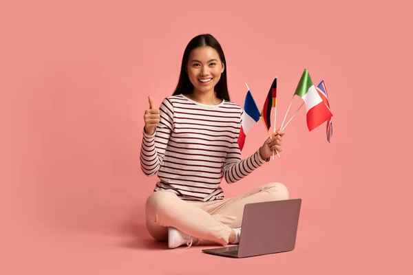 Asiatico femmina seduta con internazionale bandiere e computer portatile e mostrando pollice fino — Foto Stock
