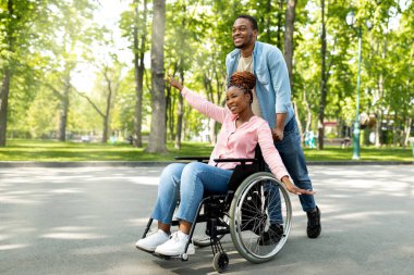 Uzun boylu, neşeli siyahi adam ve tekerlekli sandalyedeki engelli kız arkadaşı şehir parkında yürürken eğleniyorlar.