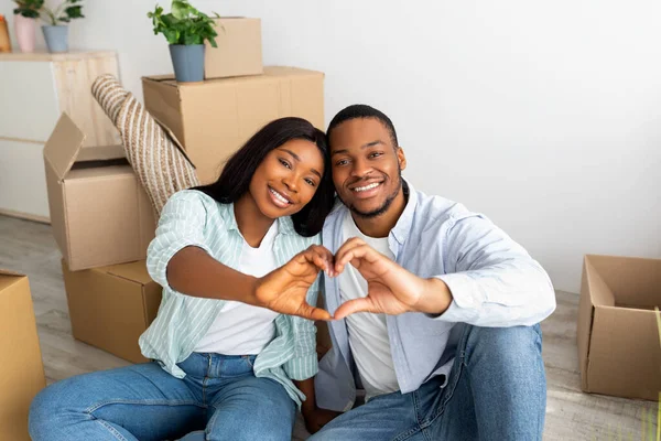 Love nest. Happy black couple sitting among moving boxes, making heart shape gesture with fingers and smiling to camera