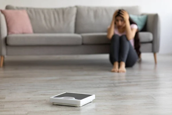 Stressed young Indian woman sitting on floor, grabbing her head in terror, looking at empty scales, feeling overweight — Stock Photo, Image