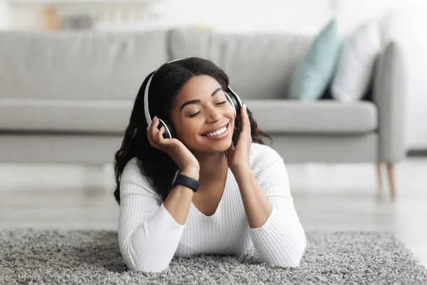 Relajada dama afroamericana escuchando música en auriculares inalámbricos mientras está acostada con los ojos cerrados en la alfombra del piso — Foto de Stock