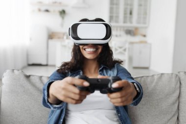 VR gözlüklü mutlu Afro-Amerikalı kadın, tutkuyla online oyunlarda oynuyor, kanepede oturuyor.