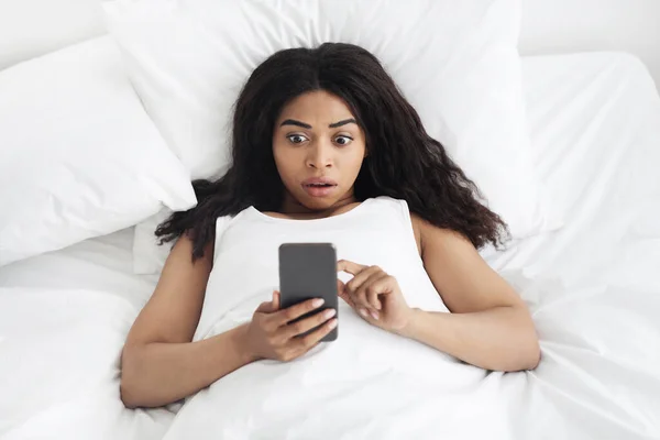 Increíbles noticias por la mañana. Mujer afroamericana conmocionada tumbada en una cama suave y mirando el teléfono inteligente, vista superior — Foto de Stock