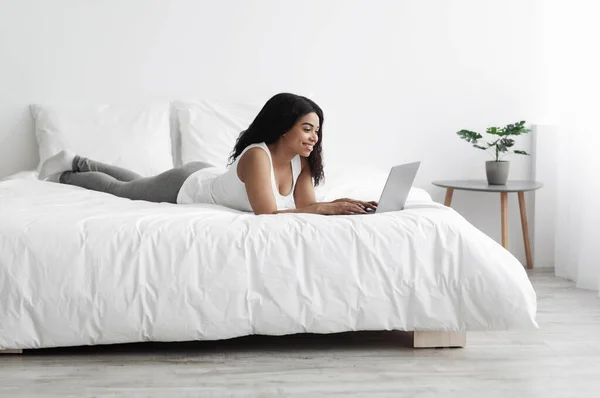 Relajada mujer negra acostada en la cama y usando un ordenador portátil, trabajando remotamente, charlando o navegando por las redes sociales — Foto de Stock