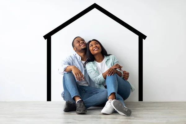 A planear a deslocalização. Sonhando casal afro-americano sentado perto da parede branca com casa desenhada e abraçando — Fotografia de Stock
