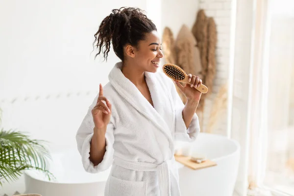 Feliz preto feminino se divertindo cantando com escova de cabelo no banheiro — Fotografia de Stock