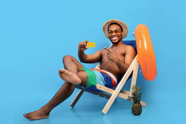 Chico negro feliz apuntando a la tarjeta de crédito mientras está sentado en el sillón en las vacaciones de verano sobre fondo azul —  Fotos de Stock