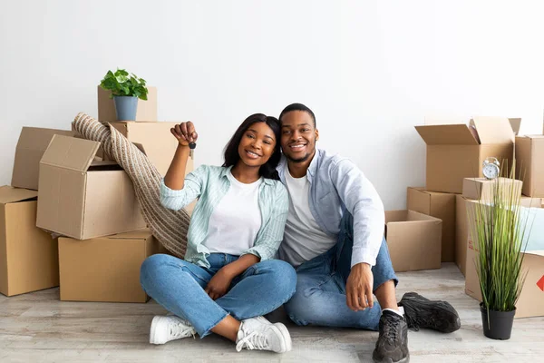 Happy african american spouses with house key and cardboard boxes looking at camera and smiling, sitting on floor — Stock Photo, Image