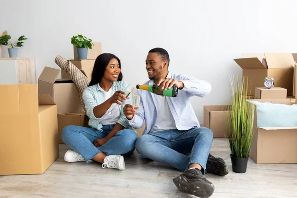 Spännande afrikanskt amerikanskt par som sitter på golvet i ny lägenhet och firar med champagne — Stockfoto