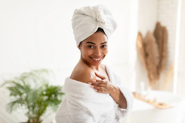 Negro mujer aplicando loción en hombro de pie en moderno cuarto de baño —  Fotos de Stock