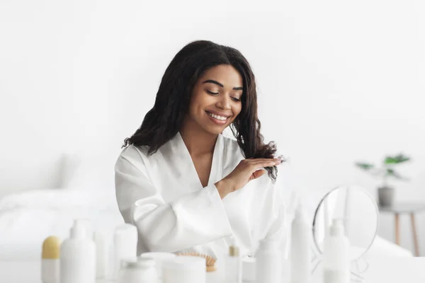 Dia de cosméticos e spa. Feliz mulher afro-americana tocando seu cabelo e olhando no espelho em casa — Fotografia de Stock