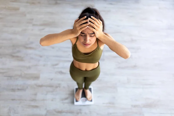 Upprörd ung indisk kvinna olycklig över sin vikt, står på skalor, hålla huvudet i besvikelse, ovan utsikt — Stockfoto