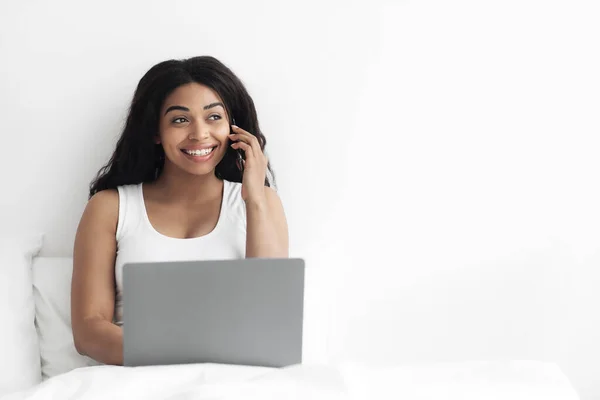 Счастливая черная женщина разговаривает по мобильному телефону и с помощью ноутбука, подтверждая свой заказ по телефону, сидя на кровати, пустое пространство — стоковое фото