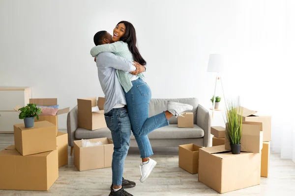 Возбужденная черная пара празднует день переезда, парень поднимает свою жену в их новой квартире, стоя среди коробок — стоковое фото