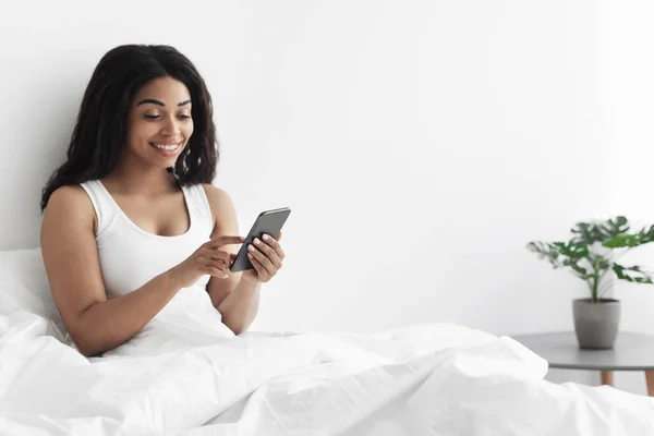 Mujer negra feliz sentado en la cama y sonriendo mientras lee un mensaje agradable en el teléfono inteligente por la mañana, espacio para copiar — Foto de Stock