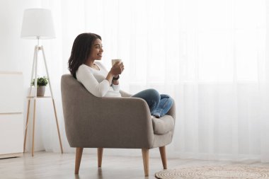 Rahatlama zamanı. Afrika kökenli Amerikalı kadın bir fincan kahveyle koltukta oturuyor ve hayal kuruyor, sabah keyfine bakıyor, boş bir alanda.
