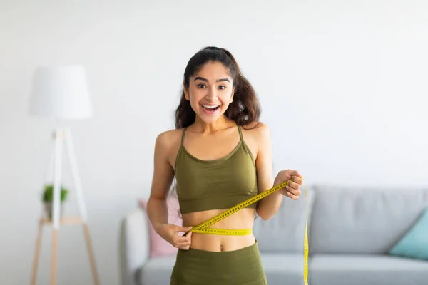 Retrato de una joven india midiendo su cintura con cinta adhesiva en el interior, sonriendo a la cámara — Foto de Stock