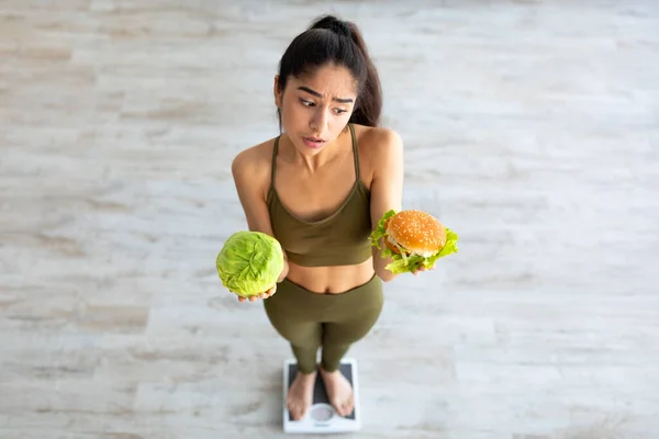 Über der Ansicht einer jungen Inderin, die Kohl und Hamburger in der Hand hält, auf einer Waage steht und ihre Mahlzeit auswählt — Stockfoto