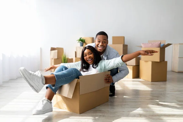 Movendo e conceito imobiliário. Joyful casal negro se divertindo e montando em caixas de papelão no novo apartamento — Fotografia de Stock