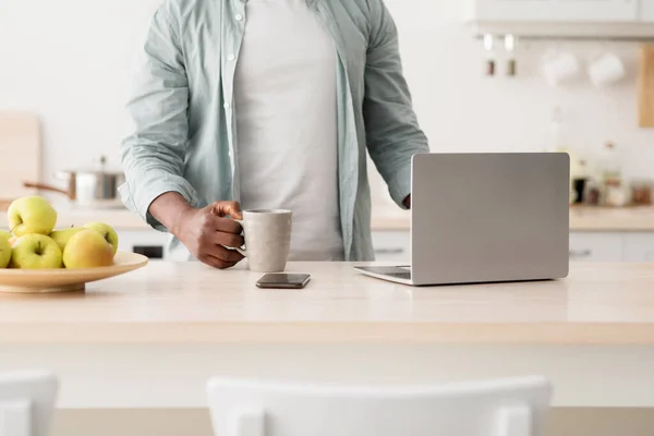Reggeli rutin fogalom. Felismerhetetlen afro-amerikai férfi laptopot használ és forró italt iszik a konyhában. — Stock Fotó