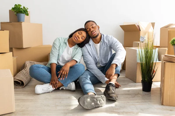 Sad black family sitting on floor with cardboard boxes around, being tired and taking break in unpacking things