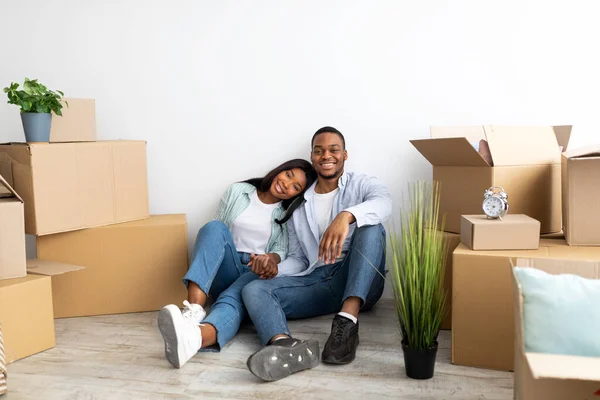 Vastgoedhuur concept. Liefdevolle Afro-Amerikaanse echtgenoten verhuizen naar een nieuw appartement, zittend tussen kartonnen dozen — Stockfoto