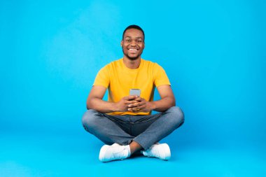 Mutlu Afrikalı Amerikalı erkek mavi arka planda oturan cep telefonunu kullanıyor.