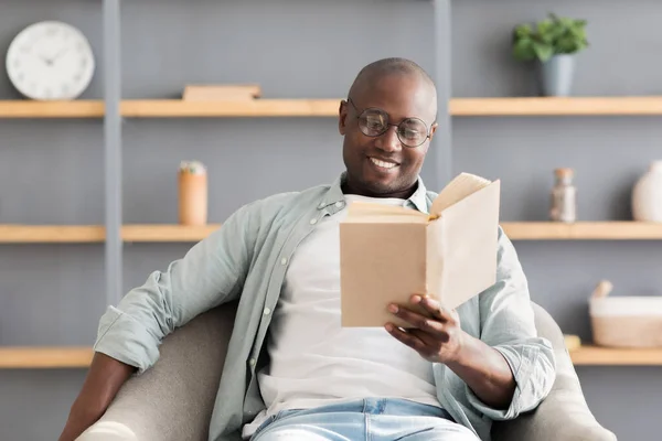 Rimani a casa a divertirti. Felice uomo maturo africano americano godendo libro di lettura, seduto in poltrona comoda, spazio libero — Foto Stock