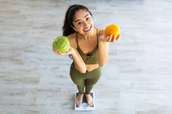 Gesunde Ernährung zur Gewichtsabnahme. Über Ansicht der Millennial Indian Lady, die Zitrusfrüchte und Gemüse hält, während sie Waagen benutzt — Stockfoto