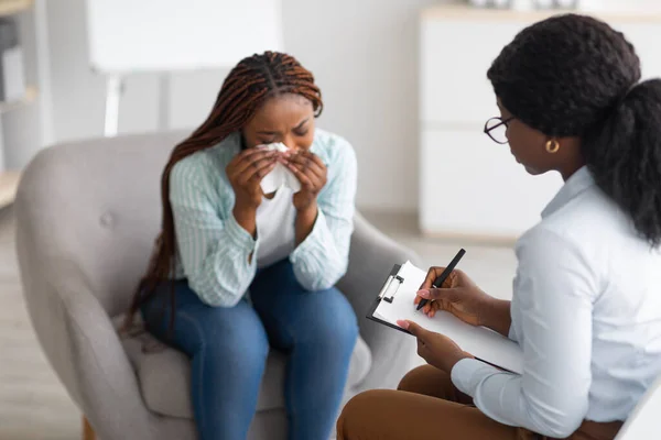 Depressieve zwarte vrouw consulterend psycholoog, zittend in een fauteuil en huilend op medisch kantoor — Stockfoto