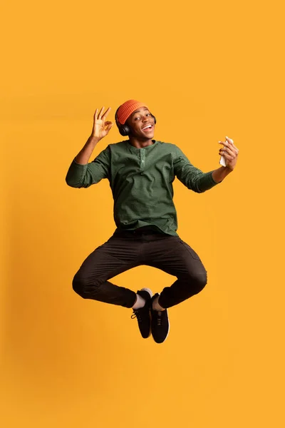 Cheerful Young Black Man In Wireless Headphones Jumping In Air With Smartphone