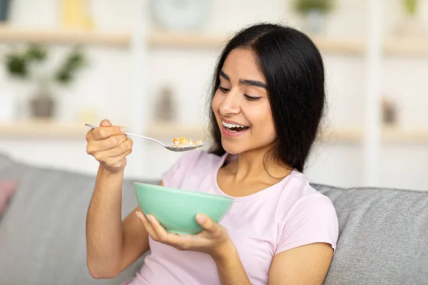 Balanserat näringskoncept. Härlig indisk dam äter läckra flingor med mjölk på mysig soffa hemma — Stockfoto