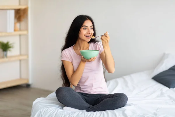 Aantrekkelijke Indiase dame eten lekker ontbijtgranen met melk terwijl zitten op comfortabel bed thuis, volledige lengte — Stockfoto