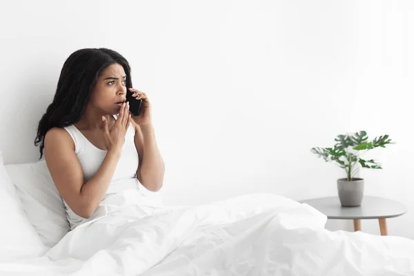 Mujer negra conmocionada sentada en la cama y hablando en el teléfono inteligente, mirando el espacio de copia, el interior del dormitorio —  Fotos de Stock