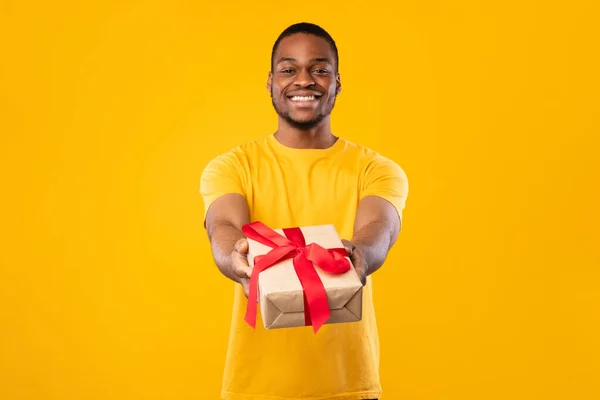 Black Guy Giving Wrapped Present Box To Camera, Yellow Background — Stock Photo, Image