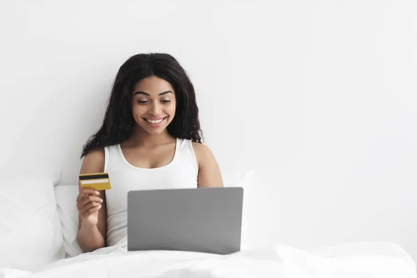 Koncepcja zakupów online. Pozytywne Afroamerykanka kobieta siedzi w łóżku z laptopem i trzyma kartę kredytową, wolne miejsce — Zdjęcie stockowe