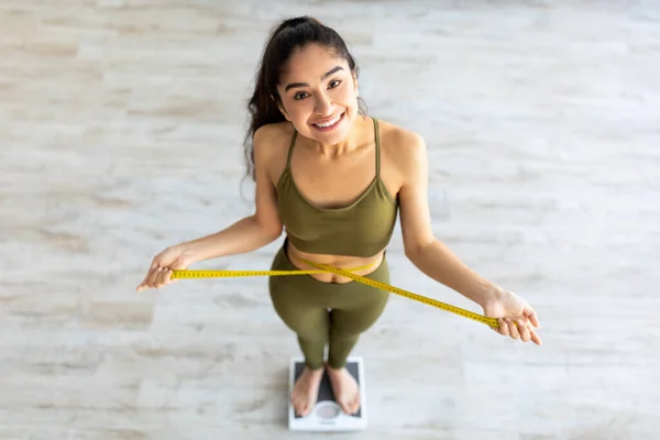 Indiano femmina di misurazione della vita mentre in piedi su scale, felice del risultato di perdita di peso di successo a casa, sopra la vista — Foto Stock