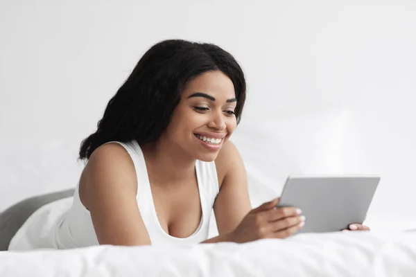Viendo video y noticias. Mujer negra emocionada sosteniendo tableta digital y acostada en la cama por la mañana, espacio libre — Foto de Stock