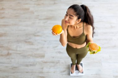 Hintli kadın sağlıksız yiyecekler yerine sağlıklı olanı seçiyor, turunçgillerin kokusunu alıyor, terazinin üzerinde dururken hamburger tutuyor.